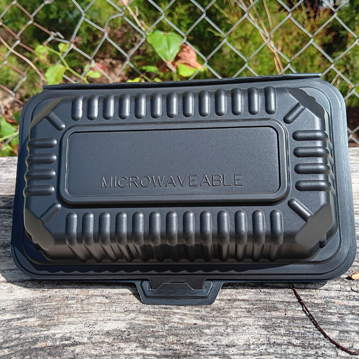 Black microwaveable food container on a wooden surface with a chain-link fence and greenery in the background.